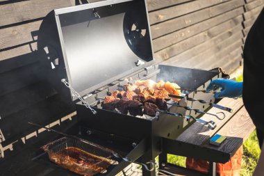 Yüzü olmayan kişi güneşli bir yaz gününde mangalda et pişiriyor. Hafta sonu pikniği. Bir kadın et pişiriyor, muhtemelen tavuk ve sığır eti, mangalın üzerinde, ahşap bir zemine kurulmuş..
