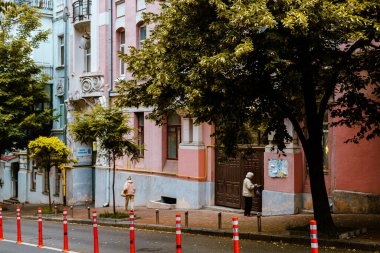 Kyiv, Ukrayna 14 Haziran 2025 Büyüleyici bir şehir caddesinde yaşlı bir kadın ahşap bir kapının yanında duruyor. Ukrayna başkentinde sakin bir şehir manzarası, pastel renkli binalar, bereketli ağaçlar ve insanların yer aldığı. 