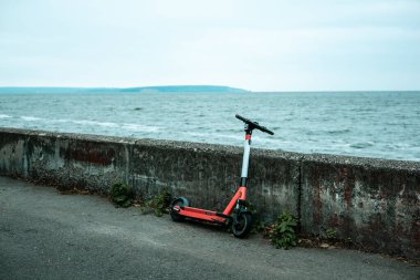 Bir elektrikli scooter bulutlu bir günde deniz kenarında oturur. Şehir manzarası sakin ve huzurlu. Bir elektrikli scooter yıpranmış bir duvara dayanır, bulutlu bir gökyüzünün altında sakin bir suya bakar.. 