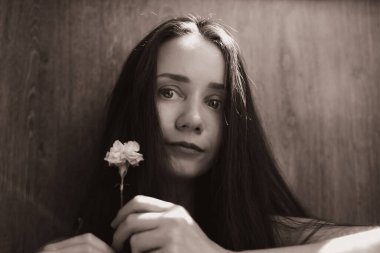 Genç bir kadın, elinde narin bir çiçek tutarak huzur içinde bakıyor. Direk kameraya bakıp hassas ve içe dönük bir atmosfer yaratan bir kızın septik tonda bir kadın portresi.