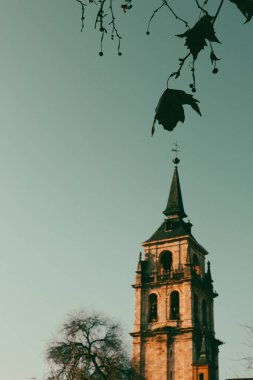 Güneşli bir günde, sonbahar yapraklarıyla eski bir kilisenin güzel kulesi dimdik ayakta duruyor. Tarihi bir kilise kulesi, ağaç dalları ve sonbahar yapraklarıyla süslenmiş pastel mavisi gökyüzüne karşı yükseliyor..
