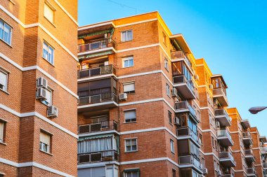 Tuğladan bir apartman dairesi açık mavi bir gökyüzünün altında, sıcak güneş ışığında duruyor. Açık mavi gökyüzünün altında tuğla cepheli, balkonlu ve klimalı bir binanın dış görüntüsü..