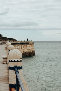 Kıyı sahnesinde, okyanusa bakan taştan bir kiremit ve balık tutan bir kişi vardı. Taştan bir balyoz, uçsuz bucaksız bir okyanus ve bulutlu bir günde balık tutan bir insanın deniz kenarındaki gezinti manzarası.