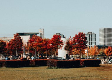 Berlin, Almanya 2 Ekim 2024 Sonbahar renkleri, modern binalar ve insanlarla dolu canlı şehir parkı sahnesi. Canlı kırmızı yapraklar ve modern mimari içeren, kentsel doğada pitoresk bir sonbahar günü.
