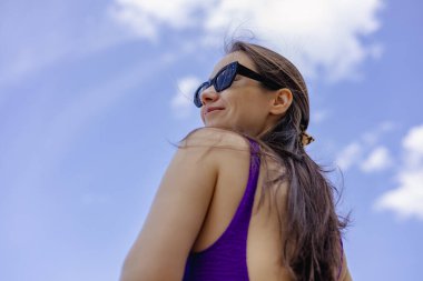 Yazın güneş gözlüğü takan bir kadın dışarıda gülümsüyor. Güneş gözlüklü, mutlu, gülümseyen bir kadın uzaklara bakar, güneş parlar. Mavi gökyüzünün altında mor bir mayo giyiyor..