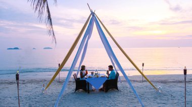 Gün batımında Koh Chang Tayland 'da bir plajda romantik bir akşam yemeği yiyen iki erkek ve kadın.