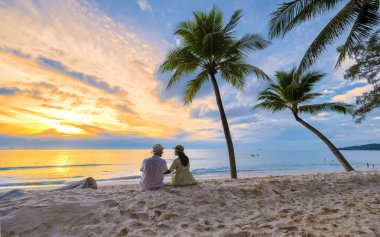 Birkaç erkek ve kadın plajda oturup kumsalda gün batımını izliyorlar. Beyaz kum ve palmiye ağaçları, Bang Tao plajı Phuket Tayland.. 