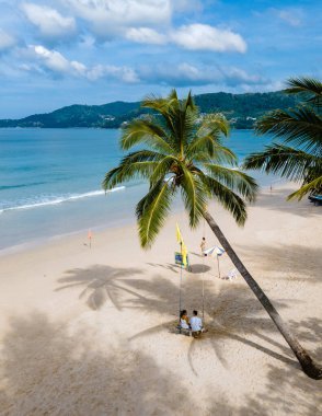 Birkaç erkek ve kadın, Patong Phuket Tayland sahilinde bir palmiye ağacının altında salıncakta sallanıyorlar, turkuaz renkli bir okyanusla. 