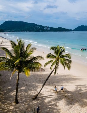 Birkaç erkek ve kadın Patong Phuket Tayland sahilinde palmiye ağacının altında salıncakta sallanıyorlar. Taylandlı kadınlar ve beyaz erkekler aşağı iniyor.