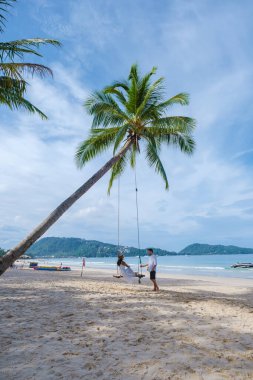 Birkaç erkek ve kadın, kasım ayının güneşli bir gününde Patong Phuket Tayland sahilinde bir palmiye ağacının altında salıncakta sallanıyorlar.
