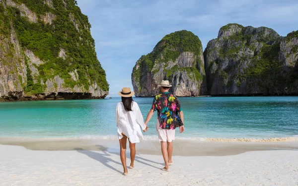 The backside of Thai women and caucasian men with a hat walk on the beach of Maya Bay, beach Koh Phi Phi Thailand in the morning with turqouse colored ocean.