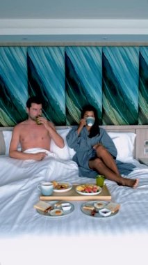 Birkaç erkek ve kadın yatakta kahvaltı yapıyor, beyaz erkekler ve Taylandlı kadınlar lüks bir otelin yatak odasında kahvaltı yapıyorlar. 