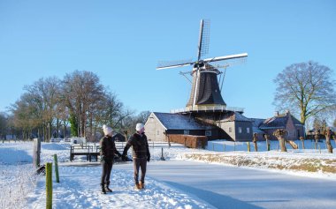 Hollanda 'da soğuk bir kış günü kar üzerinde yürüyen bir çift erkek ve kadın. Arka planda, Overijssel' de eski bir tarihi ahşap yel değirmeni var.