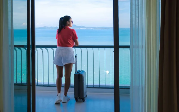 A Thai woman with hand luggage and a trolley checking in at a hotel room looking out over the ocean in Thailand.