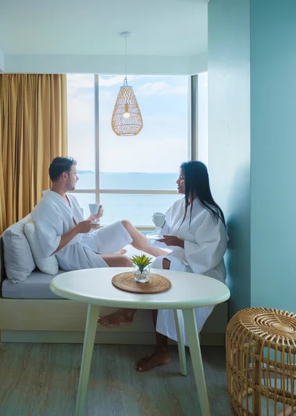 A couple of Asian women and caucasian men drink coffee in a hotel bedroom looking out over the ocean in Thailand