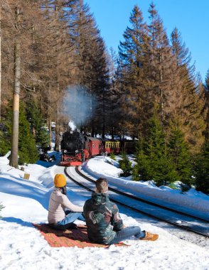 Almanya 'nın Harz milli parkında kışın buhar trenini izleyen birkaç beyaz erkek ve Asyalı kadın kışın Brocken Dağı' ndaki Buharlı Bahn treni