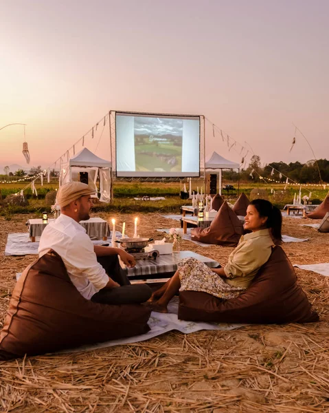 A couple of men and women watching a movie at an outdoor cinema in Northern Thailand Nan Province out over the rice paddies in Thailand, green rice fields.