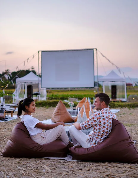 A couple of men and women watching a movie at an outdoor cinema in Northern Thailand Nan Province out over the rice paddies in Thailand, green rice fields.