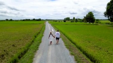 A couple walks on a road with green rice paddy fields in the countryside of Thailand.