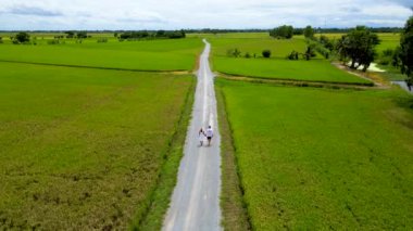 A couple walks on a road with green rice paddy fields in the countryside of Thailand.