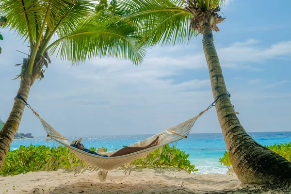 women in a hammock under a palm tree at Petite Anse beach Mahe Tropical Seychelles Islands.