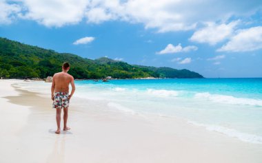 Young men in a swim short relaxing at Anse Lazio beach with turquoise colored ocean Praslin Seychelles Islands. clipart
