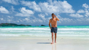 young men in swim short at a white tropical beach with turquoise colored ocean Anse Volbert beach Praslin Seychelles.