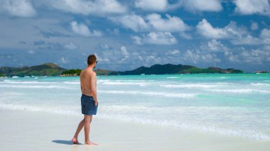 young men in swim short at a white tropical beach with turquoise colored ocean Anse Volbert beach Praslin Seychelles.