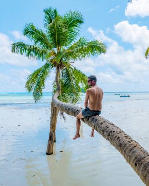 young men sitting at a palm tree on a white tropical beach with turquoise colored ocean Anse Volbert beach Praslin Seychelles.
