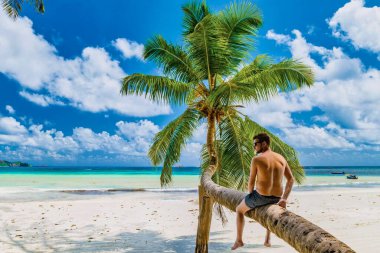 young men sitting at a palm tree on a white tropical beach with turquoise colored ocean Anse Volbert beach Praslin Seychelles.