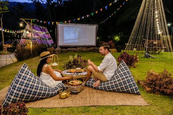 Couple men and women watching a movie in the garden of an outdoor cinema film in a tropical garden with Christmas lights. H