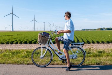 Genç adam elektrikli yeşil bisiklet bir yel değirmeni çiftliğinin yanında, yel değirmenleri, Hollanda Flevoland Noordoostpolder 'da güzel ve aydınlık bir günde izole edilmiş. 