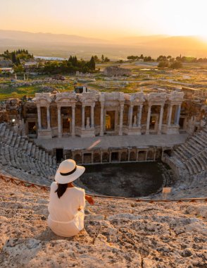 Hierapolis antik kenti Pamukkale Türkiye, Unesco harabelerinde günbatımını izleyen şapkalı genç bir kadın. Türkiye 'deki eski amfitiyatroda gün batımını izleyen Asyalı kadınlar 