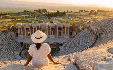 Hierapolis antik kenti Pamukkale Türkiye, Unesco harabelerinde günbatımını izleyen şapkalı genç bir kadın. Türkiye 'deki eski amfitiyatroda gün batımını izleyen Asyalı kadınlar 