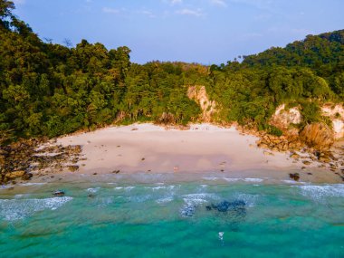 Tayland 'ın Koh Kradan adasındaki kumsalda güneş batımında palmiye ağaçlarıyla sahilde yürüyen bir çift kadın ve erkek İHA görüntüsü.