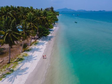 Tayland 'da Andaman Denizi' ndeki Koh Mook tropikal adasında tatilde olan bir çift kadın ve erkek, palmiye ağaçlarıyla kumsalda yürüyorlar.