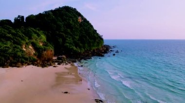 Güneşli bir günde, turkuaz renkli bir okyanusla Koh Kradan Adası sahilinde yürüyen bir çift kadın ve erkek.