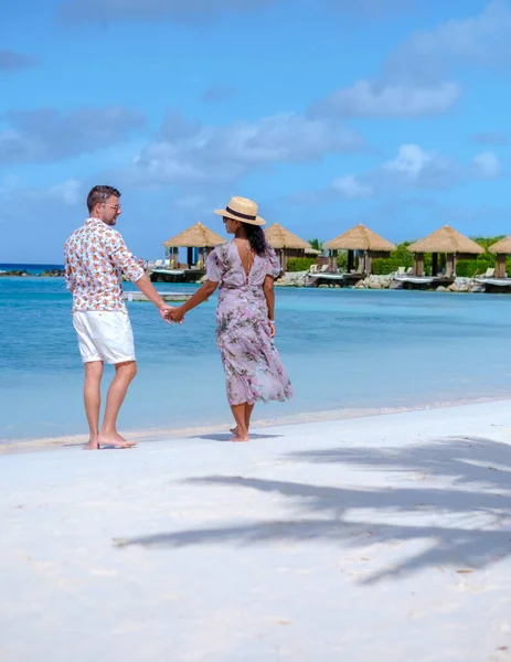 アルバ島で休暇中の男性と女性のカップルカリブ海は 豪華なリゾートでターコイズブルーの海とオーバーウォーターバンガローのヤシの木と熱帯の白いビーチを歩いています — ストック写真