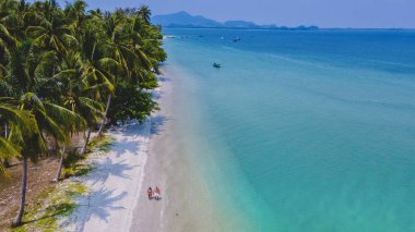 Tayland 'da Andaman Denizi' ndeki Koh Mook tropikal adasında tatil yapan bir çift kadın ve erkek palmiye ağaçlarıyla kumsalda yürüyorlar.