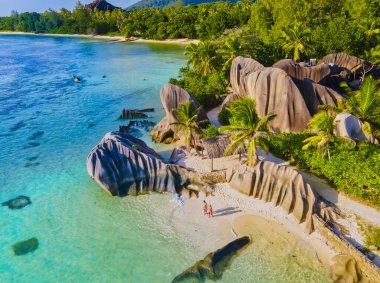 Anse Source dArgent plajı, La Digue Adası, Seyshelles, La Digue Seyşeller 'in insansız hava görüntüsü tropik adanın kuş bakışı görüntüsü, günbatımında sahilde yürüyen iki erkek ve kadın.
