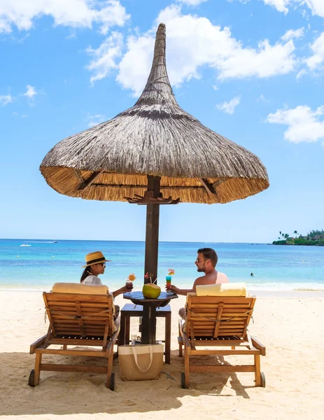 Man and Woman on a tropical beach with Chairs And Umbrella In Tropical Beach. Beautiful tranquil white sand beach with two chairs and thatched umbrella, couple drinking cocktails on the beach