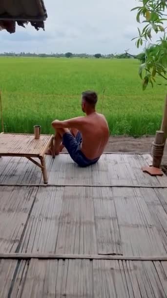 タイ中部スーパーブリ地方のグリーンライスパディフィールドと竹小屋ホームステイ農場 男性とタイの農場滞在で自分の部屋から田んぼを見渡す — ストック動画