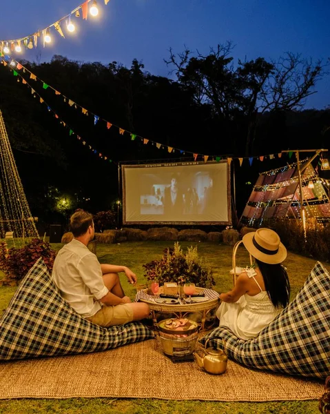 Couple men and women watching a movie in the garden of an outdoor cinema film in a tropical garden with Christmas lights. H
