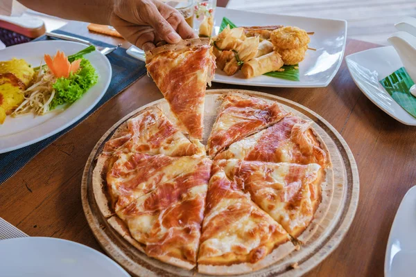 woman hand with a piece of a pizza on dinner table with pizza and Thai food on the beach in Thailand, close up of dinner table at sunset