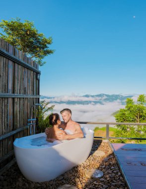 Bir çift romantik erkek ve kadın küvette tatil sırasında Chiang Rai Tayland dağlarına bakıyorlar. Dışarıda banyo, gün doğarken küvet, dağlarda sis ve sis.