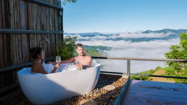 Tatil sırasında Chiang Rai Kuzey Tayland dağlarına bakan bir çift kadın ve erkek bir küvette şampanya içiyorlardı. Dışarıda banyo, gün batımında küvet.