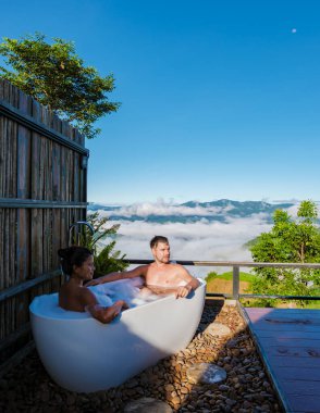 Tatil sırasında Chiang Rai Kuzey Tayland dağlarına bakan bir çift kadın ve erkek bir küvette şampanya içiyorlardı. Dışarıda banyo, gün batımında küvet.