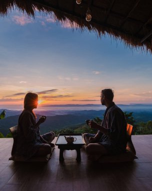 Kuzey Tayland 'da tatilde olan bir çift erkek ve kadın gün doğumunda Chiang Rai Doi Chang dağlarındaki bir kulübede kalıp ahşap balkonda çay içiyorlar.