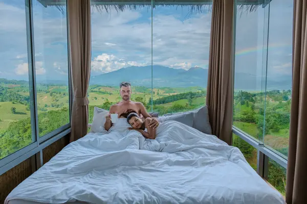 a couple of men and women laying in bed with open widows looking out over the mountains of Thailand, men and woman in bedroom with large open glass windows looking out over the green landscape