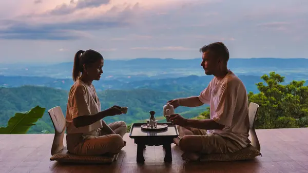 北タイでの休暇の日の出に紅茶を飲む男性と女性のカップルは 日の出に木製のバルコニーで茶を飲むチェンライドイチャン山のホームステイ小屋に滞在しています — ストック写真